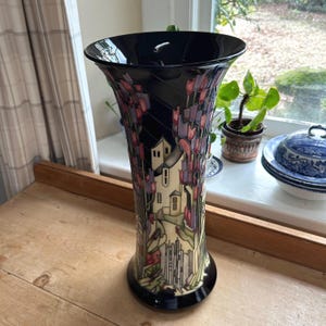 May include: A tall, black vase with a flared top and a detailed, colorful design. The vase features a landscape with buildings and floral elements in shades of purple, pink, and green. The vase is approximately 12 inches tall.