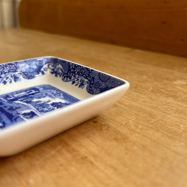 May include: A small, white, square ceramic dish with a blue and white floral pattern around the edge. The dish has a blue and white scene of a building and trees in the center.