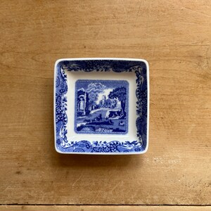 May include: A square blue and white ceramic dish with a blue and white floral border and a central image of a blue and white landscape with a bridge and a building.
