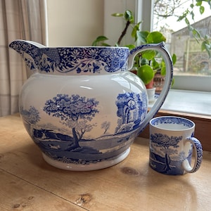Peut inclure: Un grand pichet en céramique blanche et une tasse assortie plus petite, tous deux décorés d'un motif bleu de type transfert représentant des paysages détaillés, des arbres et des ruines architecturales. Le pichet a un large bec et une anse courbée.