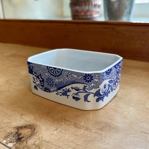 以下が含まれることがあります： 青と白のフローラルパターンが施された白いセラミック製の砂糖入れ。ボウルは長方形で、わずかに丸みを帯びたエッジがあります。