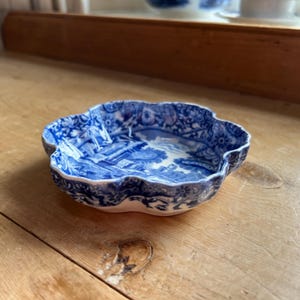May include: A small, blue and white ceramic dish with a scalloped edge. The dish features a detailed landscape scene in the center and floral patterns around the rim. The dish is sitting on a wooden surface.