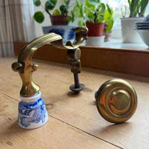 May include: A vintage brass door handle set. The set comprises a curved handle, a threaded spindle, and a round knob. The handle features a blue and white porcelain base with a decorative pattern. The brass elements show signs of age.