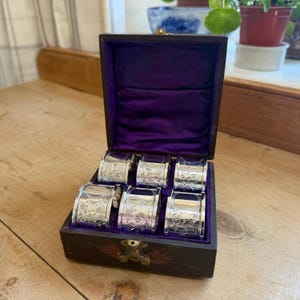 Puede incluir: Un juego de seis servilleteros plateados con grabados florales, presentados en una caja de madera vintage forrada con terciopelo morado. La caja está abierta, revelando los anillos.