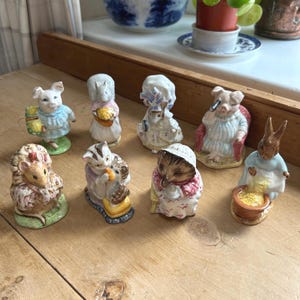 May include: A collection of ceramic figurines depicting various animals in detailed attire. The figurines include a pig, squirrel, rabbit, and hedgehog, each with unique clothing and accessories. The figurines are on a wooden surface.