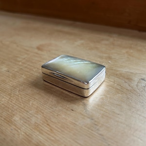 May include: A small, rectangular silver box with a pearlescent, iridescent top. The box is closed and sits on a wooden surface. The box has a hinged lid and a polished, reflective surface.