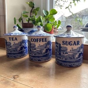 Puede incluir: Tres botes de cerámica con las etiquetas "TEA", "COFFEE" y "SUGAR". Cada bote tiene una base blanca con un diseño de paisaje azul y una tapa azul. Los botes están sobre una superficie de madera.