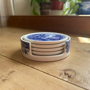 May include: A set of six ceramic coasters with a blue and white design, stacked in a white holder. The coasters feature a detailed landscape scene. The holder has a floral pattern on the sides. The coasters are on a wooden surface.