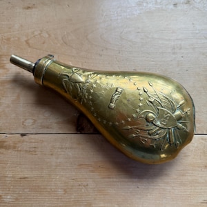 Puede incluir: Un frasco de pólvora de latón con intrincados diseños en relieve. El frasco presenta un águila, estrellas y las letras "US". Tiene un pico largo y un cuerpo redondeado, lo que sugiere un artículo histórico.