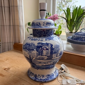 Puede incluir: Lámpara de mesa de cerámica azul y blanca con diseño de paisaje. La lámpara tiene un portalámparas en la parte superior y un cable blanco con un interruptor de encendido/apagado. La lámpara está sobre una superficie de madera.