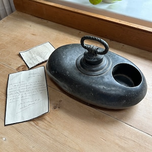 May include: An antique, oval-shaped metal hot water bottle with a handle and a circular opening. Two handwritten letters are next to the bottle. The bottle is dark gray and sits on a wooden surface.