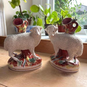 May include: Pair of ceramic sheep figurines with textured white wool, standing on a tree trunk base with colorful accents. Each sheep has a vase-like structure on its back, featuring a red and yellow design. Decorative home decor.