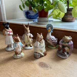 May include: A set of ceramic figurines featuring animal characters in detailed outfits. The collection includes a dog, rabbit, mouse, squirrel, duck, and frog. The figurines are painted in a variety of colours and are displayed together.