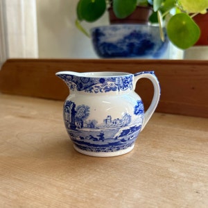 May include: A small, white ceramic creamer with a blue and white floral pattern. The creamer has a rounded body and a small spout. It is sitting on a wooden surface.