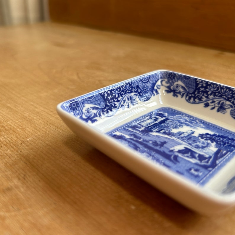 May include: A white ceramic square dish with a blue and white floral pattern around the rim. The bottom of the dish features a blue and white scene of a building and trees.