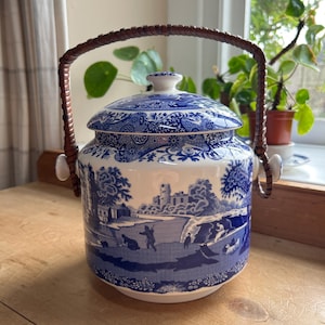 May include: A blue and white ceramic cookie jar with a brown woven handle. The jar features a traditional blue and white transferware design with a landscape scene.