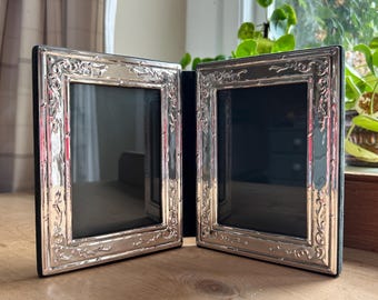 May include: Two silver-colored picture frames with ornate detailing. The frames are hinged together, creating a double-frame design. Each frame has a black backing and a clear glass or plastic front. The frames are displayed on a wooden surface.