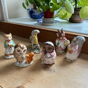 May include: A group of ceramic animal figurines, including a cat, owl, snake, hedgehog, rabbit, and squirrel. Each figure is dressed in detailed clothing. The color scheme features soft pastels and earth tones, typical of collectible figurines.