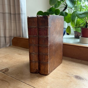 Könnte beinhalten: Zwei antike Bücher in Leder gebunden mit roten Rückenschildern und den Zahlen "1" und "2" auf den Rückenschildern. Die Bücher liegen auf einer Holzoberfläche.
