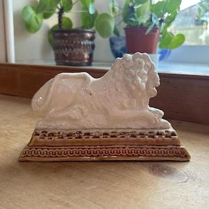 Puede incluir: Una figurita de cerámica de un león con un esmalte blanco y una base marrón. El león está acostado con la cabeza girada hacia un lado. La base tiene un patrón decorativo.