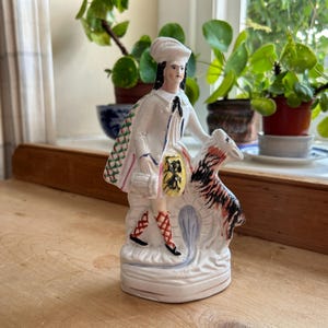 Pode incluir: Uma estatueta de cerâmica branca de uma pessoa com casaco e chapéu brancos, segurando uma bolsa e encostada em uma ovelha. A estatueta tem cabelo preto e detalhes vermelhos e pretos nas pernas. A ovelha tem marcas pretas e laranja.