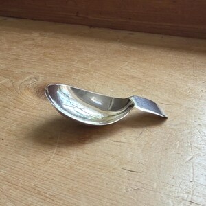 Könnte beinhalten: Ein kleiner Silberlöffel mit einer gebogenen Schüssel und einem flachen, rechteckigen Griff. Der Löffel ist hochreflektierend und zeigt die Umgebung. Der Löffel wird wahrscheinlich zum Servieren oder Essen einer kleinen Portion verwendet.