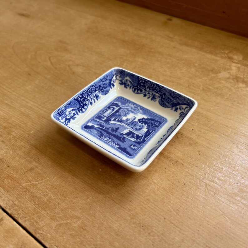 May include: A small, square, blue and white ceramic dish with a blue and white floral border and a central image of a blue and white landscape with a building and trees.
