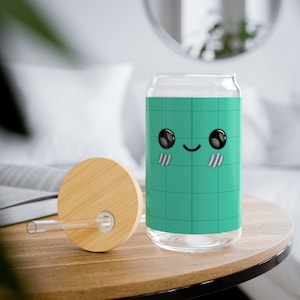 May include: A green glass can with a cute cartoon face and a bamboo lid. The can has a grid pattern and a smiling face with black eyes and pink cheeks. The lid has a hole for a straw.