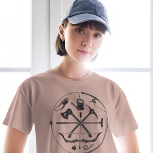 May include: A light pink t-shirt featuring a graphic of a circle containing a compass, an axe, a bow and arrow, and other camping tools. The shirt is being worn by a person wearing a blue denim baseball cap.