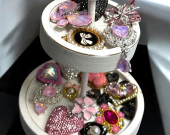 Könnte beinhalten: Ein zweistöckiges weißes Dekotablett mit verschiedenen Schmuckstücken. Der Schmuck umfasst Broschen, Anhänger und Ohrringe in Rosa-, Lila- und Schwarztönen. Einige Stücke weisen Perlen und Strasssteine auf, was eine glamouröse Präsentation ergibt.