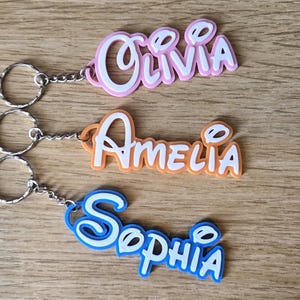 May include: Three personalised keyrings with names: Olivia in pink, Amelia in orange, and Sophia in blue. Each keyring has a silver ring and chain. The names are in a playful, stylised font, ideal for gifts or personal use.