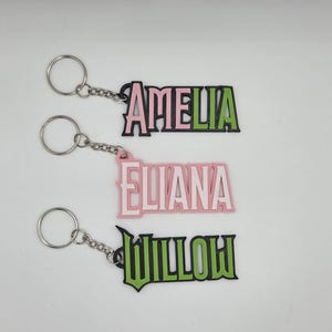 May include: Three personalised name keyrings. The keyring for "Amelia" has a black outline with pink and green lettering. The "Eliana" keyring is pink with a white outline. The "Willow" keyring has a green outline with green lettering.