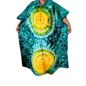 May include: A floor-length kaftan featuring a tie-dye pattern with two large, circular yellow sunburst designs centered on a deep teal and dark green background. The garment has wide, loose sleeves and a round neckline adorned with a beaded necklace.