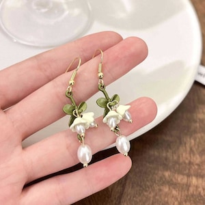 May include: A pair of dangle earrings featuring a floral design. The earrings have gold-colored hooks, green stems, and white bell-shaped flowers with pearl-like accents. Each earring has a dangling, teardrop-shaped pearl.