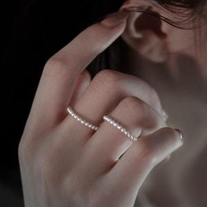 May include: Two silver rings with small pearl-like beads. The rings are worn on the index and middle fingers of a hand.