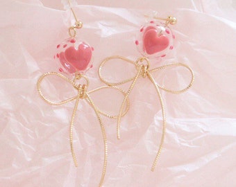 May include: A pair of gold earrings with a pink heart charm inside a clear glass bubble and a gold wire bow. The earrings are on a pink background.