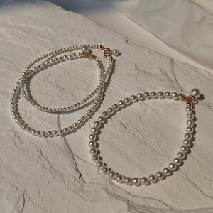May include: Two pearl necklaces with gold clasps on a white textured surface. The necklaces are made of small, white pearls.