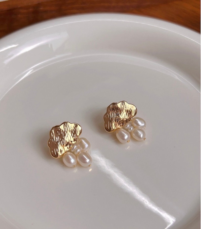 Puede incluir: Un par de pendientes de nube de oro con detalles de perlas. Cada pendiente presenta una nube de oro texturizada con un racimo de peque&ntilde;as perlas blancas que cuelgan de la parte inferior.
