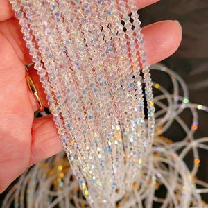 May include: Strands of iridescent, clear crystal beads. The beads are small, faceted, and strung together, creating a shimmering effect. The crystals reflect light, displaying a rainbow of colors. The beads are held in a hand, with a gold ring visible.
