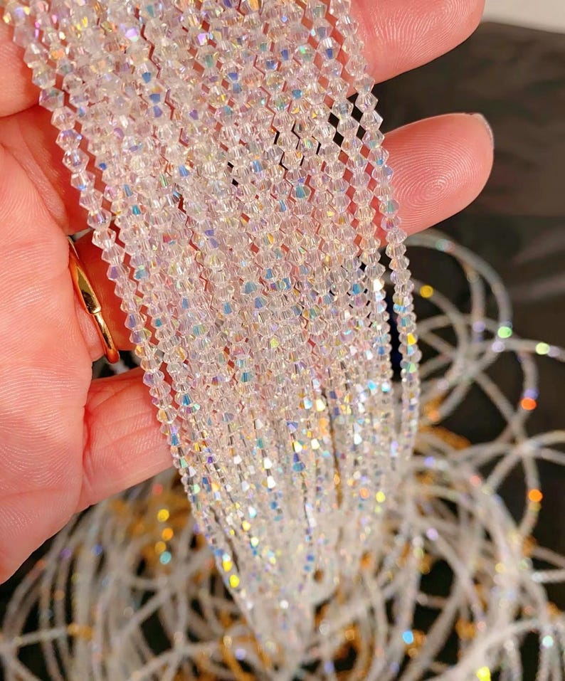May include: Strands of iridescent, clear crystal beads. The beads are small, faceted, and strung together, creating a shimmering effect. The crystals reflect light, displaying a rainbow of colors. The beads are held in a hand, with a gold ring visible.