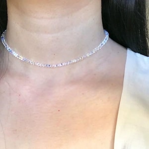 May include: A delicate necklace composed of small, clear beads with subtle blue and white accents. The necklace is worn around the neck, resting on the skin. The background is a soft, neutral tone, highlighting the jewelry.