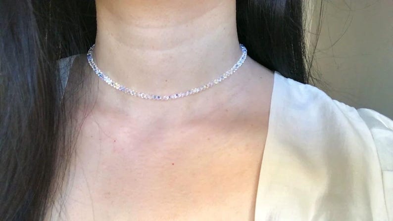 May include: A delicate necklace composed of small, clear beads with subtle blue and white accents. The necklace is worn around the neck, resting on the skin. The background is a soft, neutral tone, highlighting the jewelry.