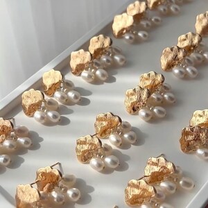Puede incluir: Un par de pendientes de tono dorado con un dise&ntilde;o en forma de nube y detalles de perlas. Cada pendiente presenta una forma de nube texturizada con tres perlas colgantes.
