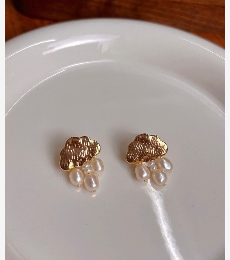 Puede incluir: Un par de pendientes de nube de oro con gotas de perlas. Los pendientes est&aacute;n sobre una superficie blanca.