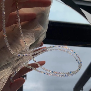 May include: A delicate, clear crystal necklace with iridescent beads. The necklace is composed of multiple strands of small, round crystals, creating a shimmering effect. The clasp is gold-toned. The necklace is displayed against a soft, white fabric.