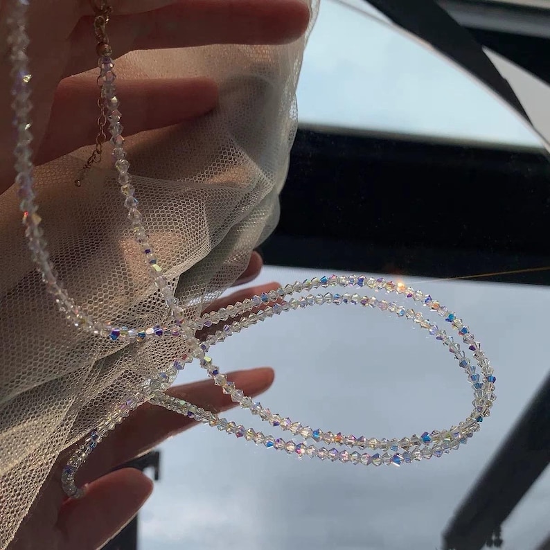 May include: A delicate, clear crystal necklace with iridescent beads. The necklace is composed of multiple strands of small, round crystals, creating a shimmering effect. The clasp is gold-toned. The necklace is displayed against a soft, white fabric.