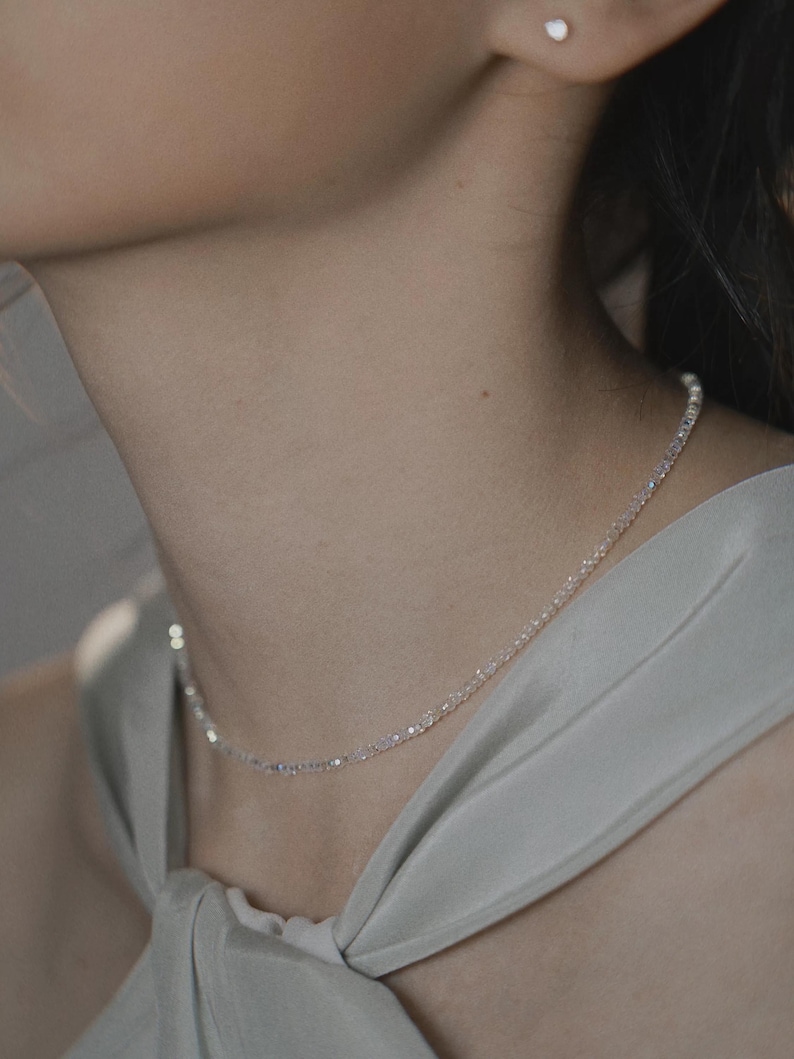 May include: A delicate, shimmering necklace with small, evenly spaced beads. The necklace is worn around the neck, resting on the collarbone. The image also shows a small, round earring and a light-colored top.