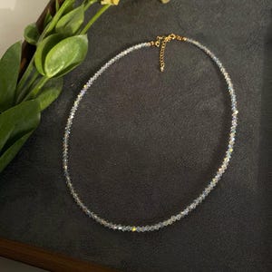 May include: A delicate necklace featuring small, iridescent beads. The necklace has a gold-colored clasp and chain. A yellow flower and green leaves are in the background, set against a dark surface.