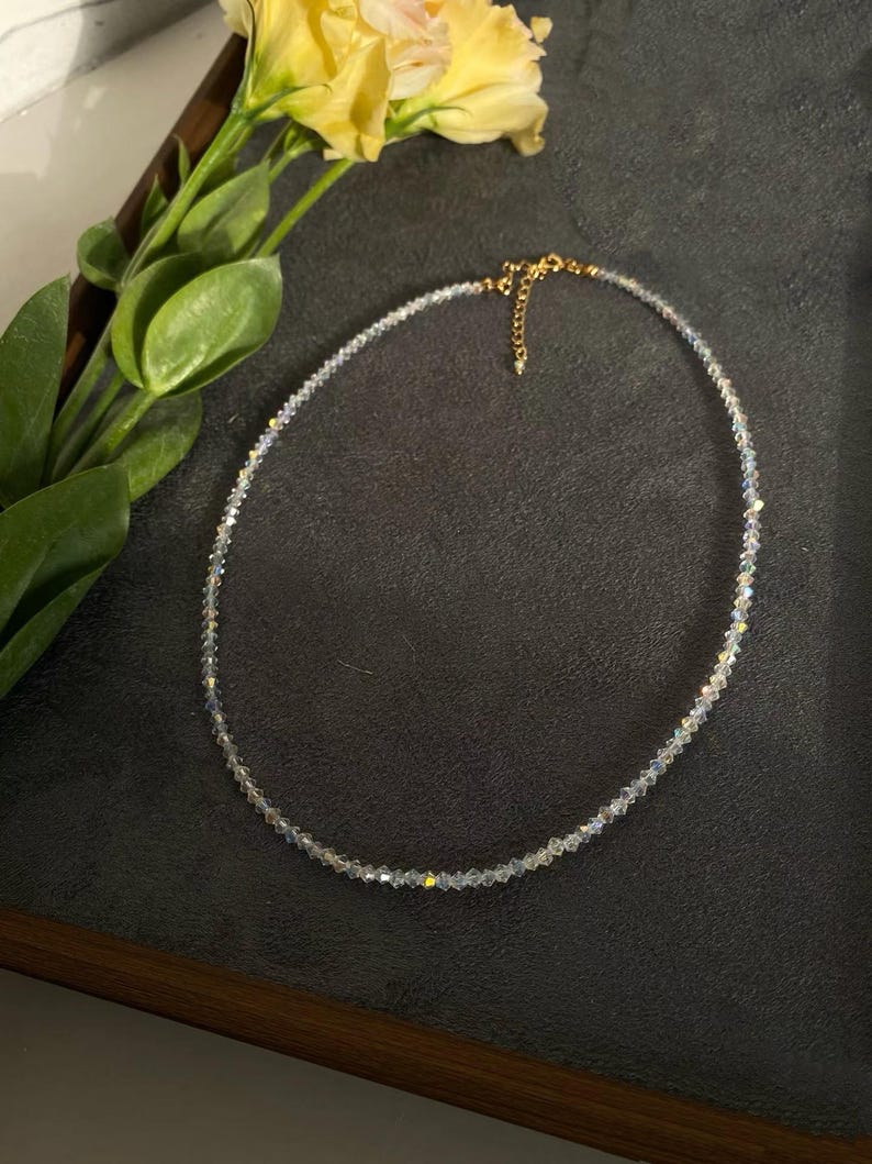 May include: A delicate necklace featuring small, iridescent beads. The necklace has a gold-colored clasp and chain. A yellow flower and green leaves are in the background, set against a dark surface.