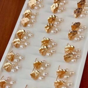 Puede incluir: Una muestra de pendientes de tono dorado con detalles de perlas. Cada pendiente presenta un dise&ntilde;o texturizado en forma de nube con tres perlas colgantes.